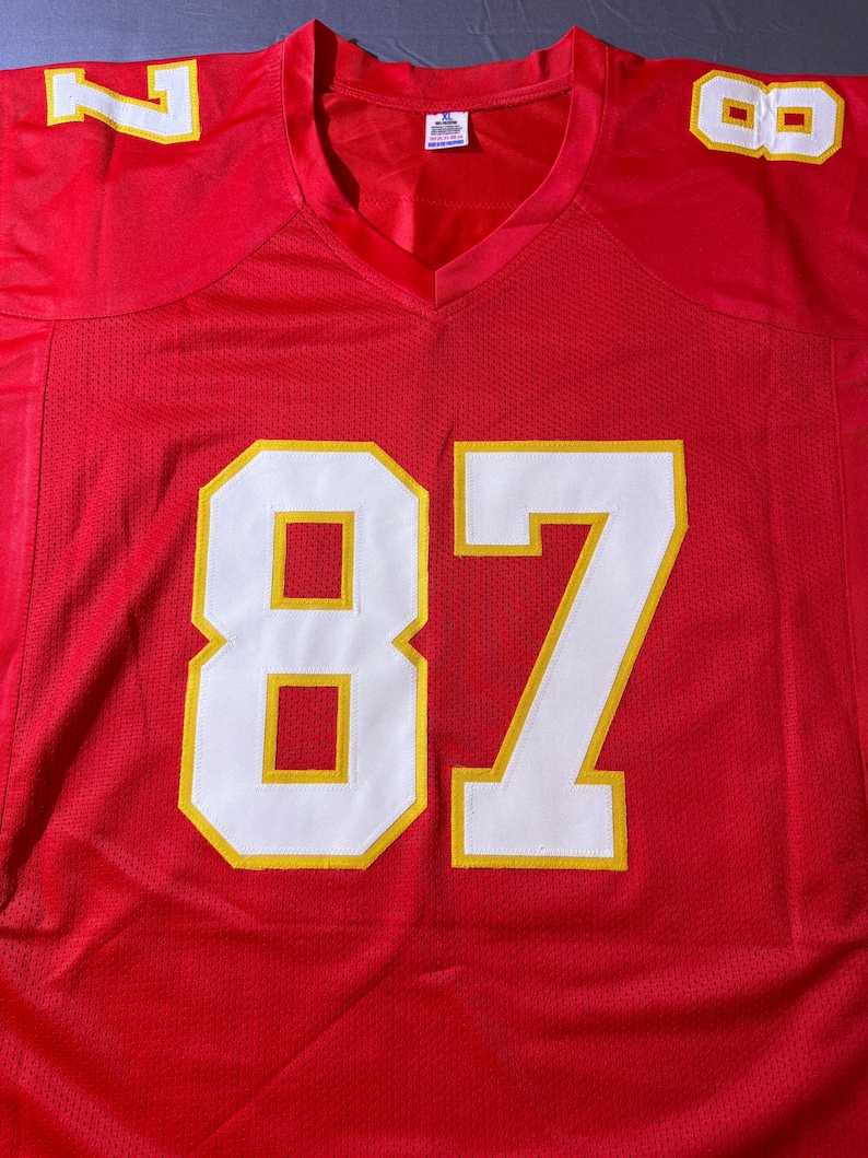 May include: A red football jersey with white and yellow accents. The jersey features the number 87 in large white numerals with yellow outlines on the front. The numbers 7 and 8 are also visible on the shoulders. The jersey has a V-neck.