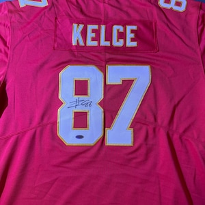 Op de afbeelding: Een rood voetbalshirt met de naam "KELCE" en het nummer "87" in wit met gouden rand. Het shirt heeft ook de nummers "1" en "8" op de schouders. Het shirt lijkt gesigneerd te zijn.