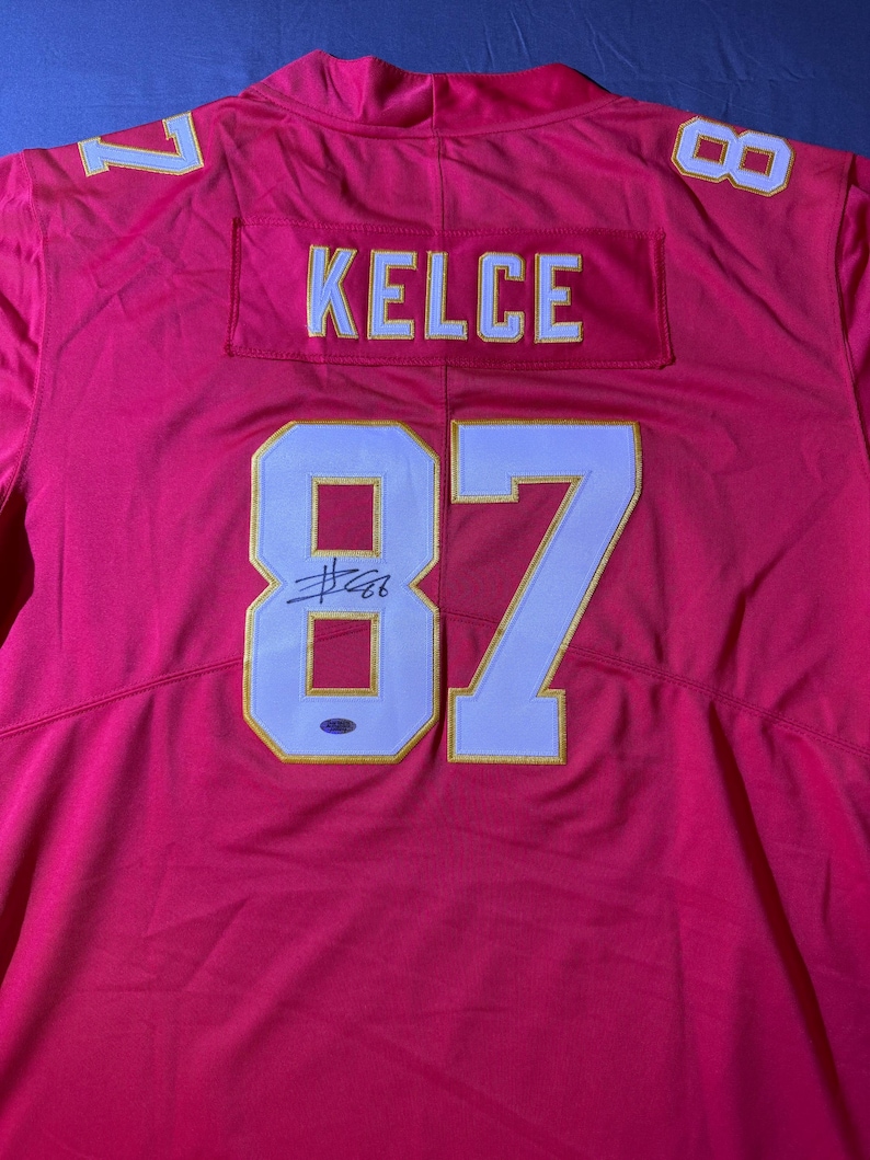 Op de afbeelding: Een rood voetbalshirt met de naam "KELCE" en het nummer "87" in wit met gouden rand. Het shirt heeft ook de nummers "1" en "8" op de schouders. Het shirt lijkt gesigneerd te zijn.