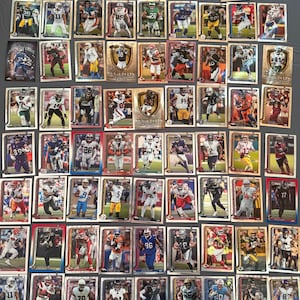 Puede incluir: Una colección de cromos de fútbol americano con varios jugadores en poses de acción. Las tarjetas muestran colores vibrantes y logotipos de equipos, algunas tarjetas están resaltadas con detalles de lámina dorada. Las tarjetas están dispuestas sobre una superficie gris.