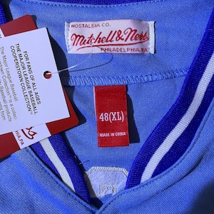 May include: A light blue baseball jersey with a royal blue and white striped collar. The interior features a Mitchell & Ness Nostalgia Co. label, a red tag marked 48(XL) Made in China, and a white hang tag with Major League Baseball text.