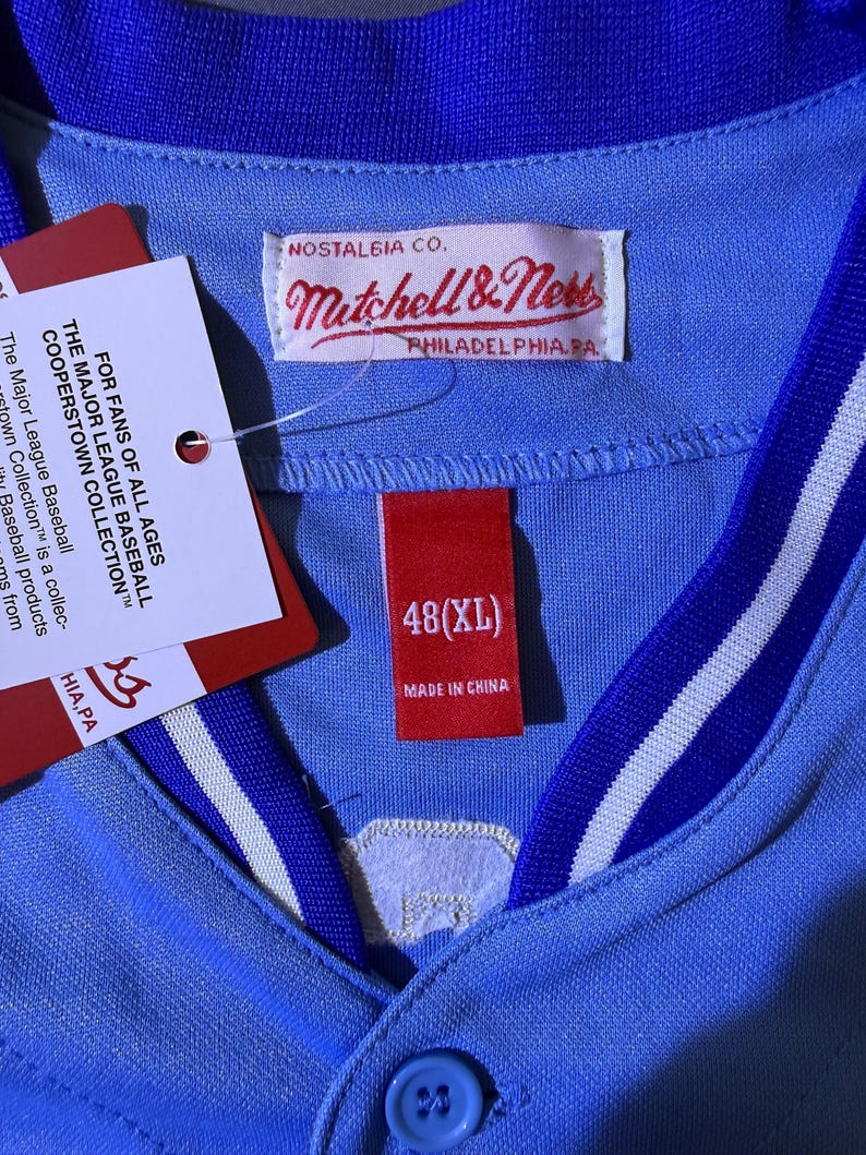 May include: A light blue baseball jersey with a royal blue and white striped collar. The interior features a Mitchell & Ness Nostalgia Co. label, a red tag marked 48(XL) Made in China, and a white hang tag with Major League Baseball text.