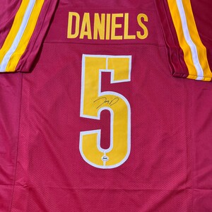May include: A red football jersey with the name "DANIELS" in yellow block letters across the shoulders and the number "5" in yellow on the front. The sleeves have yellow and white stripes.