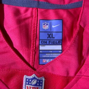 Op de afbeelding: Een felroze NFL-shirt met een mesh onderkant. Het shirt heeft het NFL-logo en een Nike-logo. Het maatlabel is XL. Gemaakt in Honduras.