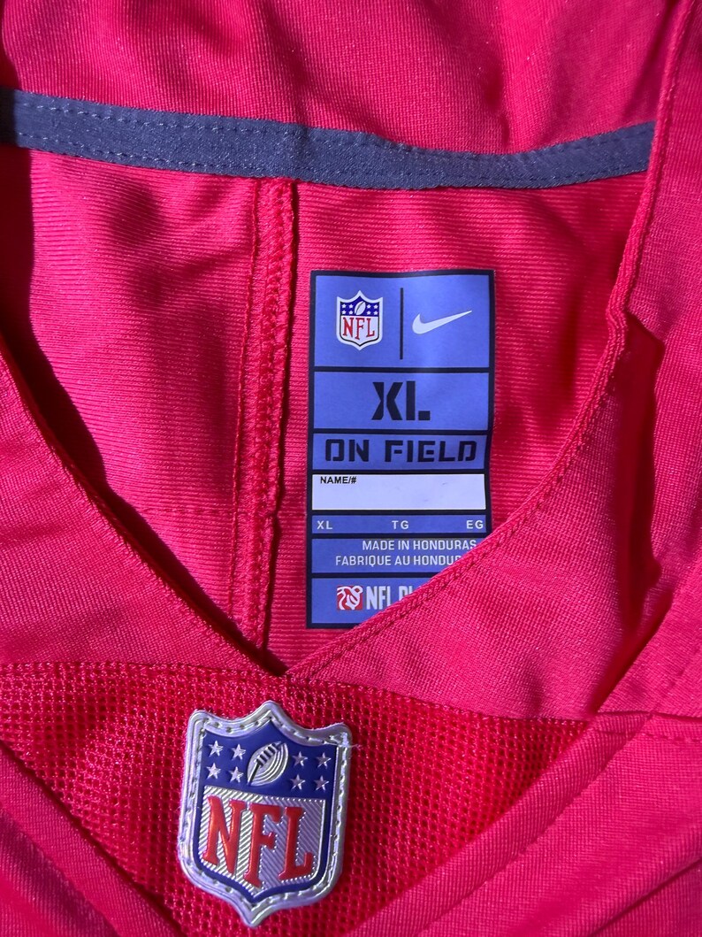 Op de afbeelding: Een felroze NFL-shirt met een mesh onderkant. Het shirt heeft het NFL-logo en een Nike-logo. Het maatlabel is XL. Gemaakt in Honduras.