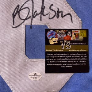 May include: A light blue fabric surface featuring a large white embroidered letter with a black ink autograph across it. A rectangular black certificate of authenticity card and a small oval white security sticker with the text VS A53322 are present.