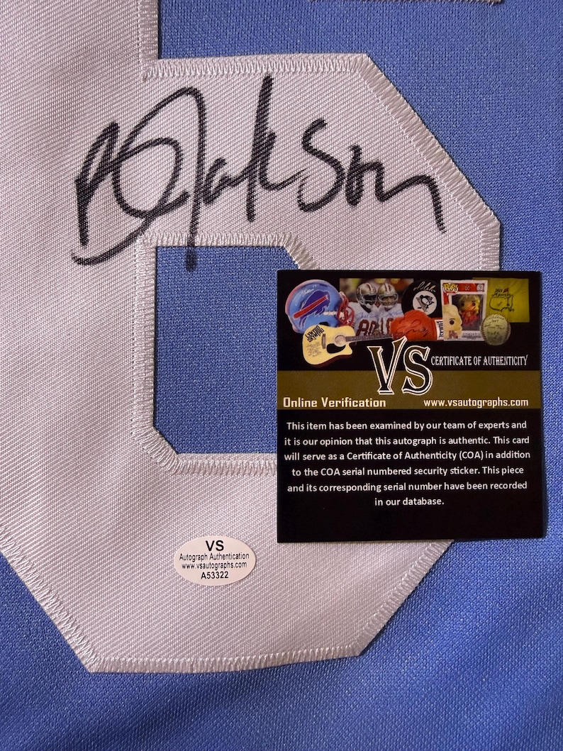 May include: A light blue fabric surface featuring a large white embroidered letter with a black ink autograph across it. A rectangular black certificate of authenticity card and a small oval white security sticker with the text VS A53322 are present.