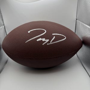 May include: A brown American football with a white signature. The football has a textured surface. The signature is in a cursive style. The football is oval-shaped.