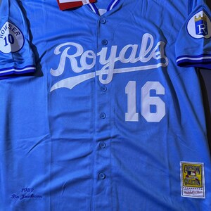 May include: A light blue button-down baseball jersey featuring the white script text Royals and the number 16 on the front. The sleeves have patches with the number 10 and a crown logo. It includes a Cooperstown Collection tag and 1987 Bo Jackson text.
