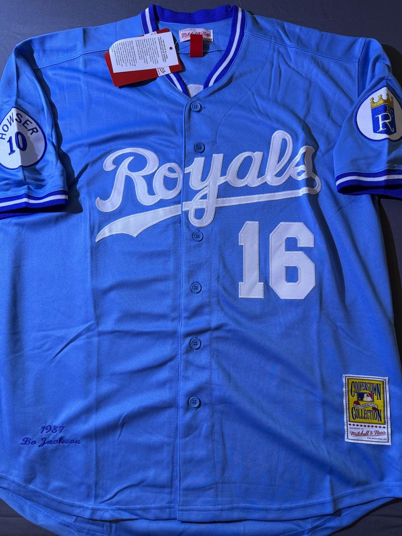 May include: A light blue button-down baseball jersey featuring the white script text Royals and the number 16 on the front. The sleeves have patches with the number 10 and a crown logo. It includes a Cooperstown Collection tag and 1987 Bo Jackson text.
