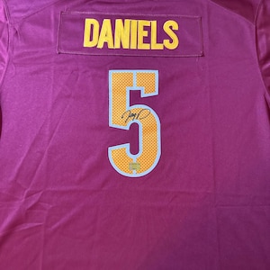 Pode incluir: Uma camisola desportiva bordeaux com o nome DANIELS em letras amarelas sobre o número 5. O número 5 é amarelo com um contorno azul claro e um padrão de pontos, e inclui uma assinatura preta e um pequeno autocolante holográfico.