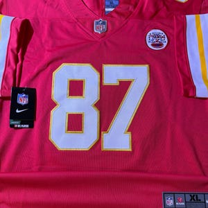 Op de afbeelding: Een rood voetbalshirt met het nummer 87 in wit en goud. Het shirt heeft het NFL-logo en een Nike-label. Het shirt is maat XL.