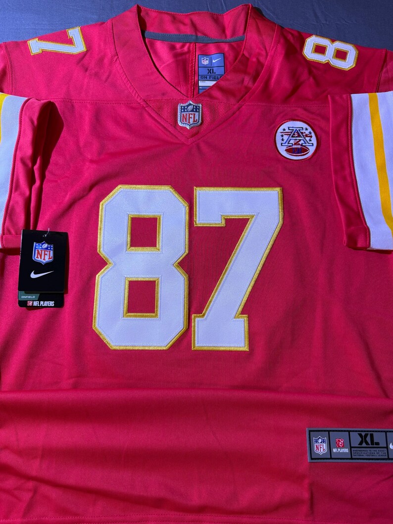 Op de afbeelding: Een rood voetbalshirt met het nummer 87 in wit en goud. Het shirt heeft het NFL-logo en een Nike-label. Het shirt is maat XL.
