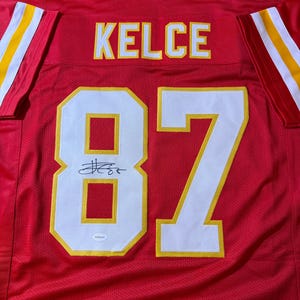 May include: A red football jersey with the name "KELCE" and the number "87" in white and yellow. The jersey has a signature above the number. The sleeves have yellow and white stripes.