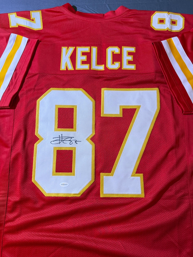 May include: A red football jersey with the name "KELCE" and the number "87" in white and yellow. The jersey has a signature above the number. The sleeves have yellow and white stripes.