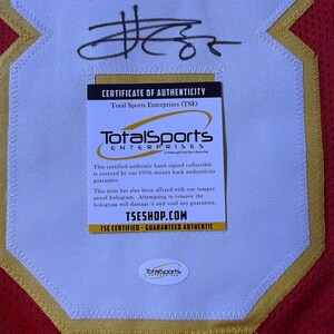 May include: A red and gold sports jersey with a white patch featuring a black signature and a "Certificate of Authenticity" from Total Sports Enterprises. The certificate guarantees authenticity and includes the website TSESHOP.COM.
