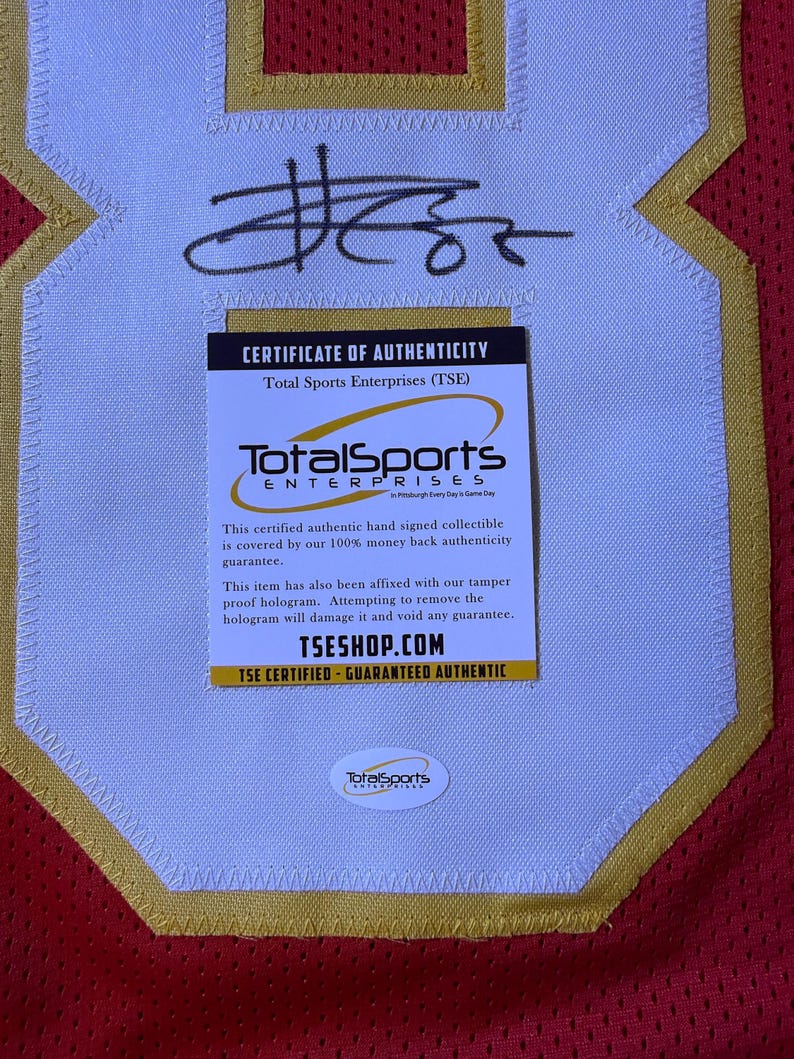 May include: A red and gold sports jersey with a white patch featuring a black signature and a "Certificate of Authenticity" from Total Sports Enterprises. The certificate guarantees authenticity and includes the website TSESHOP.COM.