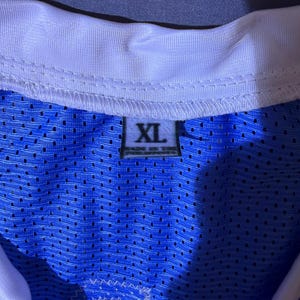 May include: A close-up of a blue mesh jersey with a white collar. The jersey has an XL size tag. The fabric is a vibrant blue with small perforations. The collar is white with a stitched design.