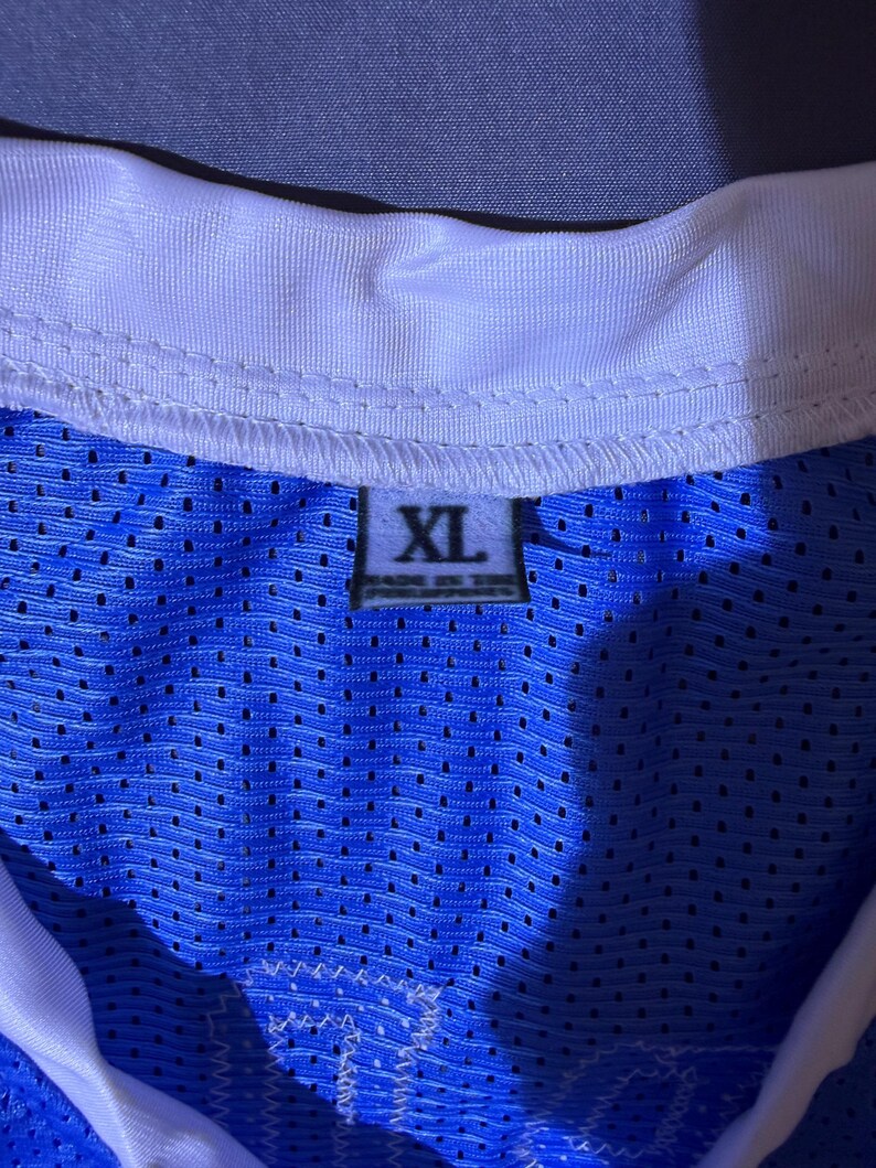 May include: A close-up of a blue mesh jersey with a white collar. The jersey has an XL size tag. The fabric is a vibrant blue with small perforations. The collar is white with a stitched design.