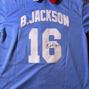 May include: A light blue sports jersey featuring the name B. JACKSON and the number 16 in white lettering on the back. The number 16 includes a black signature. Attached to the collar are two rectangular product tags with Mitchell & Ness branding.