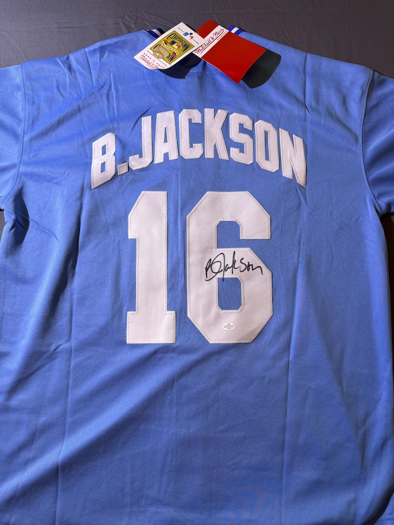 May include: A light blue sports jersey featuring the name B. JACKSON and the number 16 in white lettering on the back. The number 16 includes a black signature. Attached to the collar are two rectangular product tags with Mitchell & Ness branding.