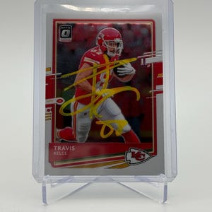 Puede incluir: Una tarjeta coleccionable deportiva con un jugador de fútbol americano con uniforme rojo, sosteniendo un balón. La tarjeta tiene un diseño rojo y plateado con el texto "Optic" y el nombre del jugador, "Travis Kelce". La tarjeta está en un soporte de plástico transparente.