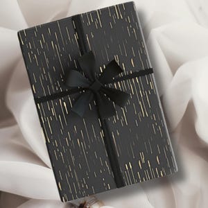 May include: A black gift box with a black ribbon and bow. The box is wrapped in black paper with gold stripes.