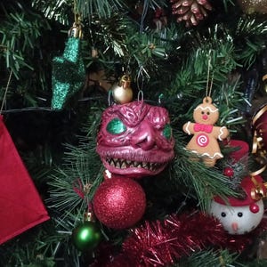 May include: A decorated Christmas tree featuring a variety of ornaments. A pink, monster-faced bauble with green eyes is near a red glitter ball and a gingerbread man decoration. Other ornaments include a green star and a snowman.