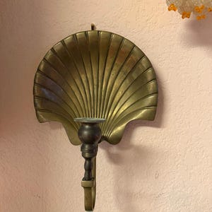 May include: A vintage brass wall sconce in the shape of a scallop shell. The sconce has a dark brown candle holder and a hook for hanging. The shell design features radiating lines, and the brass has a warm, aged patina.