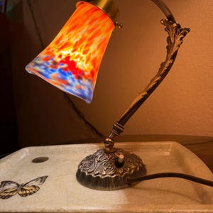 May include: A decorative table lamp with a bronze finish. The lamp features a colorful, cone-shaped shade with orange, red, and blue hues. The base has an ornate, floral design. The lamp is turned on, illuminating the colorful shade.