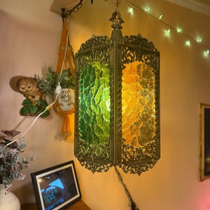 May include: A hanging pendant lamp featuring a rectangular brass frame with ornate filigree detailing. The lamp has textured glass panels in alternating shades of green and amber, suspended from a brass chain and ceiling mount fixture.