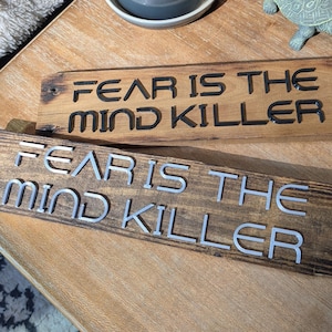 May include: Two wooden signs with the phrase "FEAR IS THE MIND KILLER" in a bold font. One sign has black lettering, and the other has silver lettering. The signs are on a wooden surface.