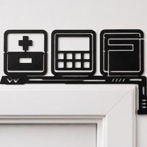 May include: A black metal door corner topper featuring three square icons: a medical cross and minus sign, a grid pattern, and a stylized letter F. The design includes a horizontal bar with geometric cutouts that rests along the door frame edge.
