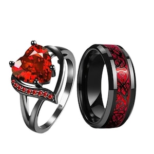 May include: A pair of black wedding bands with red accents. The woman's ring features a large heart-shaped red gemstone. The man's ring has a red and black design.