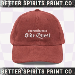May include: A brick-red corduroy baseball cap with the text "currently on a Side Quest" embroidered in white. The cap has a curved brim and a rounded top. The words "BETTER SPIRITS PRINT CO." are printed at the top and bottom of the image.