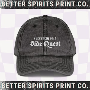 Könnte beinhalten: Verwaschene schwarze Baseballkappe mit dem weißen Text "currently on a Side Quest". Die Kappe hat einen gebogenen Schirm und einen Verschluss mit Stoffband. Der Text "BETTER SPIRITS PRINT CO." ist oben und unten im Bild.