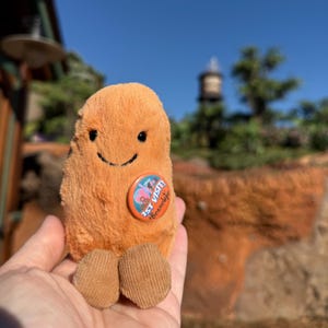 May include: A plush, orange bean-shaped toy with a smiling face and corduroy feet. It has a circular pin that reads "1st Visit! Beanelope". The toy is held in front of a blurred outdoor background with a blue sky.