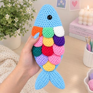 May include: A crochet fish toy featuring a light blue body with a single black eye and colourful, overlapping scales in orange, green, white, purple, yellow, pink, dark green, red, and lavender. The fish has a distinct tail and rounded fins.