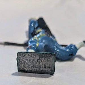 Puede incluir: Un soldado de juguete pintado de azul y blanco con un arma y una base que dice "TAINS LTD. PRO. PRIDTOYS LONDON, ENGLAND."