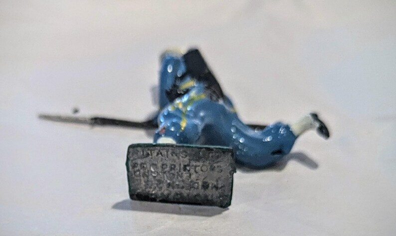 Puede incluir: Un soldado de juguete pintado de azul y blanco con un arma y una base que dice "TAINS LTD. PRO. PRIDTOYS LONDON, ENGLAND."