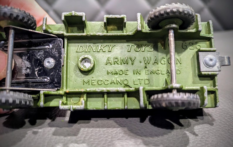 Puede incluir: Un carro de juguete verde del ej&eacute;rcito con ruedas negras y un eje de metal. La parte inferior del carro tiene el texto "Dinky Toys Army Wagon Made in England Meccano Ltd 625" impreso en &eacute;l.