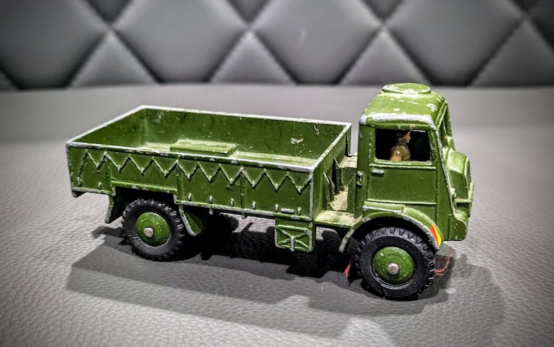 Puede incluir: Un cami&oacute;n de juguete verde con una plataforma y una figura de conductor en la cabina. El cami&oacute;n tiene un dise&ntilde;o detallado con una superficie texturizada y una franja blanca en el lateral.