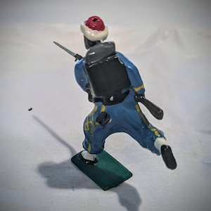 Puede incluir: Una figurita de soldado de juguete pintada de azul y amarillo con un sombrero rojo y blanco, una mochila negra y un rifle. El soldado est&aacute; corriendo y est&aacute; de pie sobre una base verde.