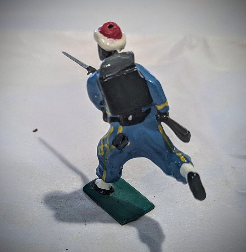 Puede incluir: Una figurita de soldado de juguete pintada de azul y amarillo con un sombrero rojo y blanco, una mochila negra y un rifle. El soldado est&aacute; corriendo y est&aacute; de pie sobre una base verde.