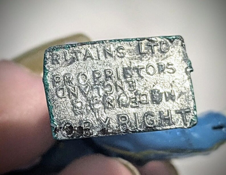 Puede incluir: Primer plano de una etiqueta de metal plateado con el texto "Britains LTD Proprietors of the "Britains" Registered Trade Mark Copyright".