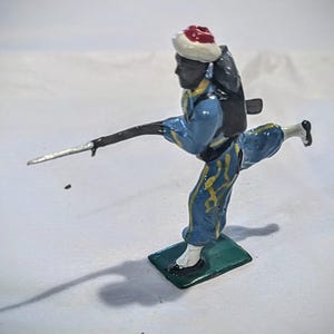 Puede incluir: Una figurita de soldado de juguete pintada de azul y amarillo con un sombrero rojo y blanco, sosteniendo una lanza plateada y llevando una mochila negra. El soldado est&aacute; de pie sobre una base verde.