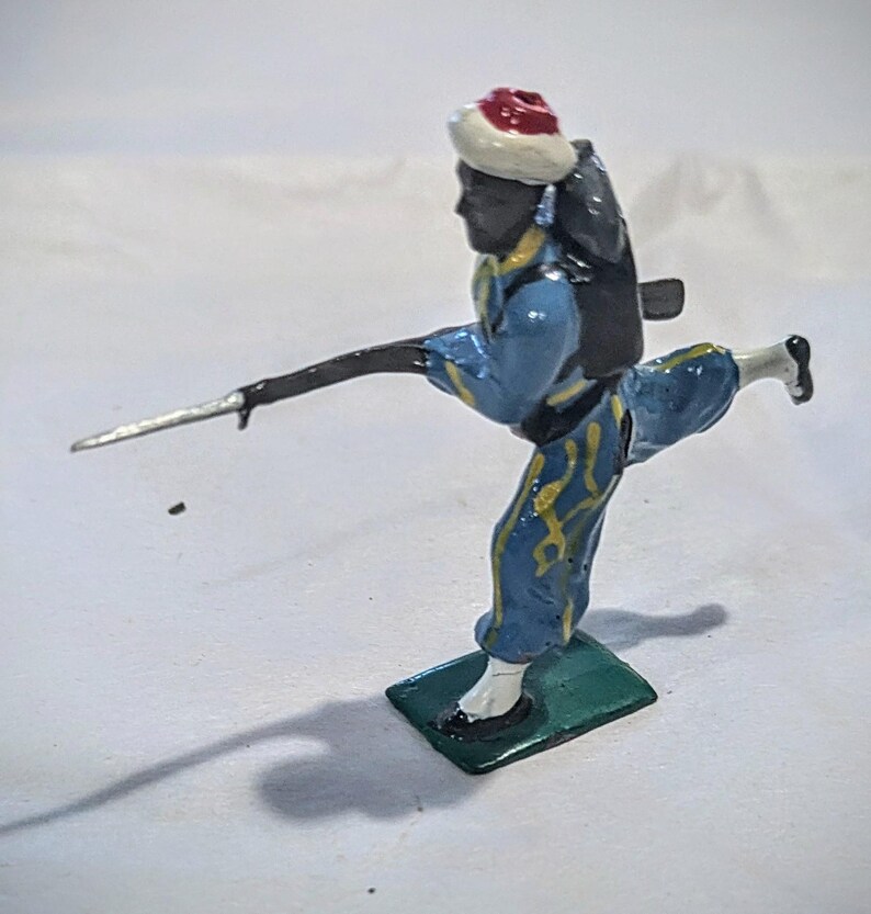 Puede incluir: Una figurita de soldado de juguete pintada de azul y amarillo con un sombrero rojo y blanco, sosteniendo una lanza plateada y llevando una mochila negra. El soldado est&aacute; de pie sobre una base verde.