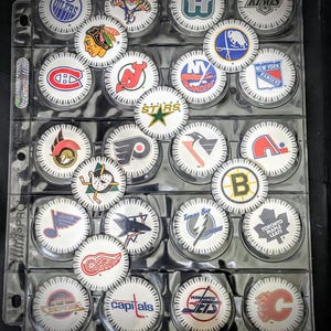 May include: A collection of 24 hockey team logo pucks in a clear plastic binder. The pucks are arranged in rows of four. The logos include the Oilers, Blackhawks, Jets, Kings, Rangers, Islanders, Devils, Stars, Flyers, Penguins, Bruins, Maple Leafs, Canadiens, Capitals, Canucks, Red Wings, Lightning, and more.