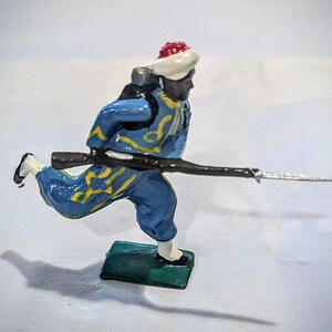 Puede incluir: Una figurita de soldado de juguete pintada de azul y amarillo corriendo con un rifle. El soldado lleva un sombrero blanco y rojo y tiene la cara negra.