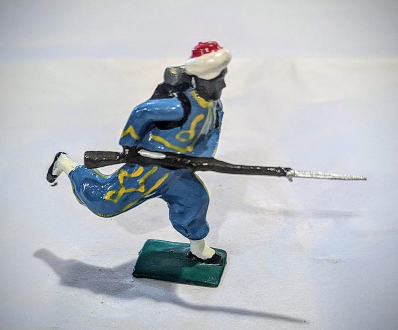 Puede incluir: Una figurita de soldado de juguete pintada de azul y amarillo corriendo con un rifle. El soldado lleva un sombrero blanco y rojo y tiene la cara negra.